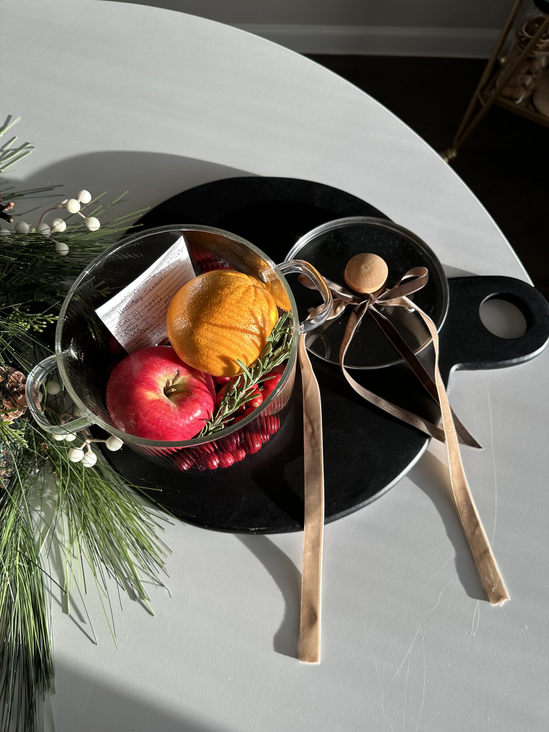 Clear pot with apple, cranberry and oranges with greenery on a black platter